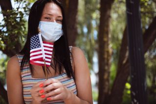 Crop anonymous lady in face mask looking at camera while standing with colorful American flag on Independence Day in park