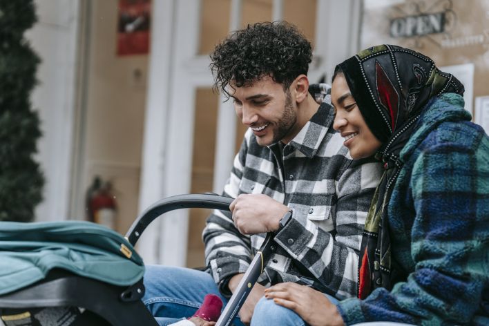 Cheerful young multiethnic spouses smiling and looking at baby in stroller